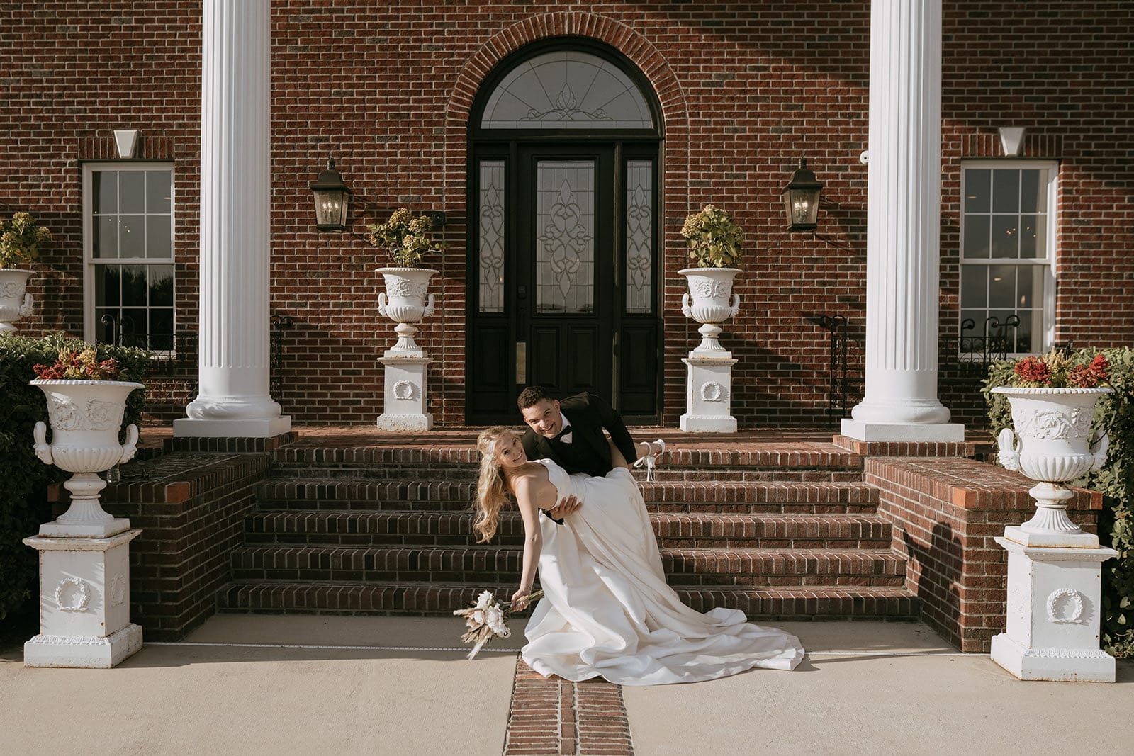 Wesley and Jessica Cameron on their own wedding day, dipping mid-dance on the brick steps of a columned venue.